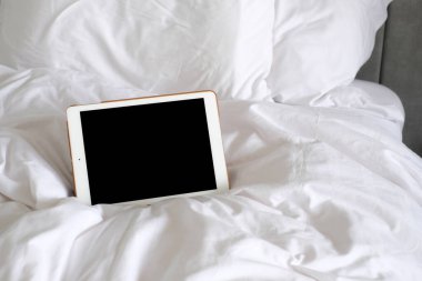 Yatak odasında beyaz battaniye ve yastıkla yatağın üzerindeki tablet boş ekran, uykusundan önce sosyal medyayı kullanıyor.