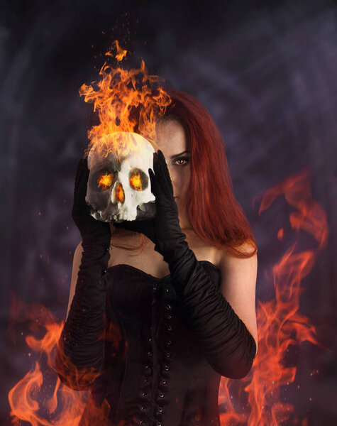 A beautiful redheaded girl in black corset and gloves holding a burning human skull.