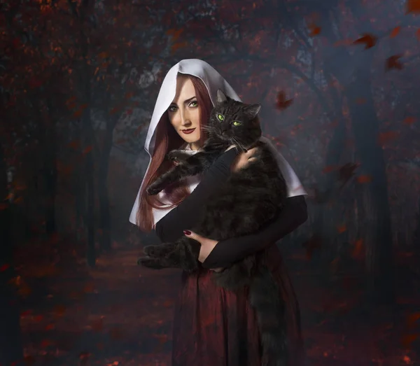 Ormanda bir kara kedi ayakta tutan beyaz kukuletalı pelerin Kızıl saçlı güzel genç kız.