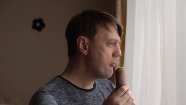 Um homem come um gelado à janela a olhar através de cortinas brilhantes. — Vídeo de Stock