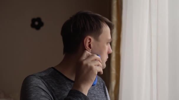 A man shaves standing in front of a window, pushing back a white curtain — Stock Video