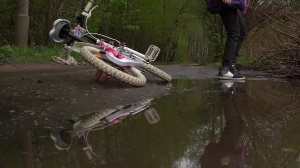 Menina lava os pés em uma poça, com uma bicicleta por uma estrada da floresta no parque — Vídeo de Stock