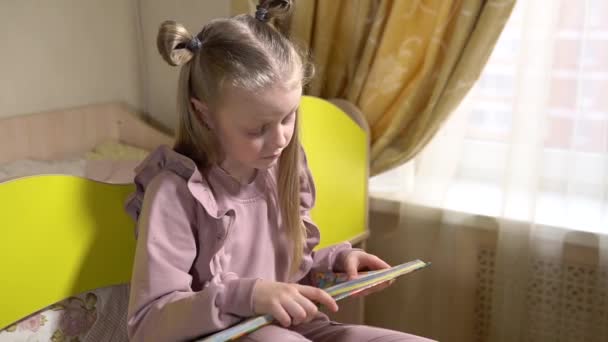 Vidám olvasás egy könyvet ül az ágyon egy padláson egy örömteli arcát egy kislány egy fényes szobában otthoni ruhákban. Egy fényes ablakhoz.. — Stock videók
