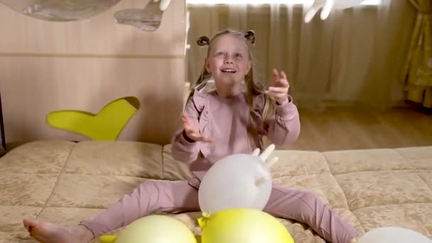 행복 한 한 소녀가 노란 장갑 과 흰색 풍선이 달린 부드러운 소파에 있는 아름다운 집에서 풍선 장갑을 끼고 노는 모습 — 비디오