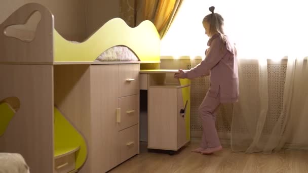 Menina alegre muda a mesa de móveis de baixo da cama, mudando o sistema modular de móveis em uma casa grande, um berço com grandes paredes amarelas. Em uma sala de luz — Vídeo de Stock