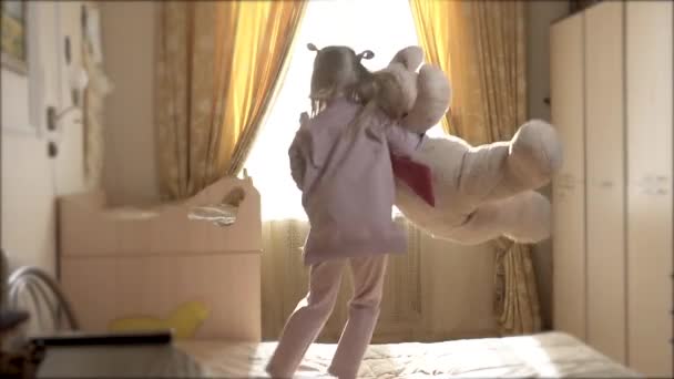 A menina brinca com ursinho pulando na cama durante o dia em casa. Feliz e alegre — Vídeo de Stock