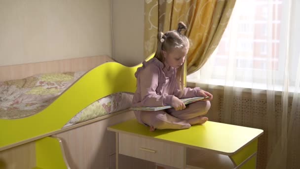 Happy lee un libro sentado en las escaleras sobre ruedas con una niña alegre en la habitación de los niños con ropa de moda. En contra de una ventana soleada . — Vídeos de Stock