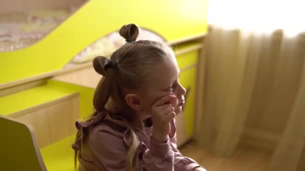 En sorgsen ung flicka sitter i ett trapphus på vinden sängen och är ledsen och tänker igen nära den gula spjälsängen, mot bakgrund av det soliga fönstret — Stockvideo