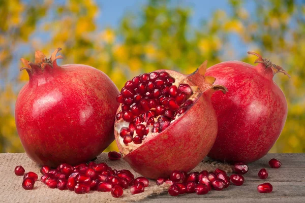 Zwei ganze Granatäpfel und die Hälfte auf dem alten Holzbrett mit verschwommenem Gartenhintergrund — Stockfoto