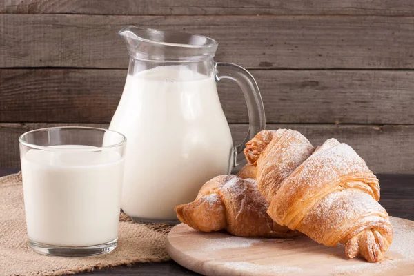 Kancsó és pohár tej, croissant-t, egy fából készült háttér — Stock Fotó
