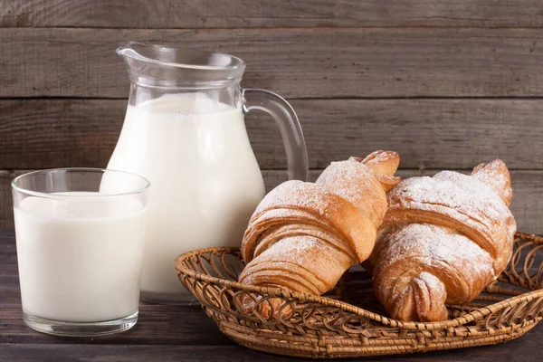 Kancsó és pohár tej, croissant-t, egy fából készült háttér — Stock Fotó