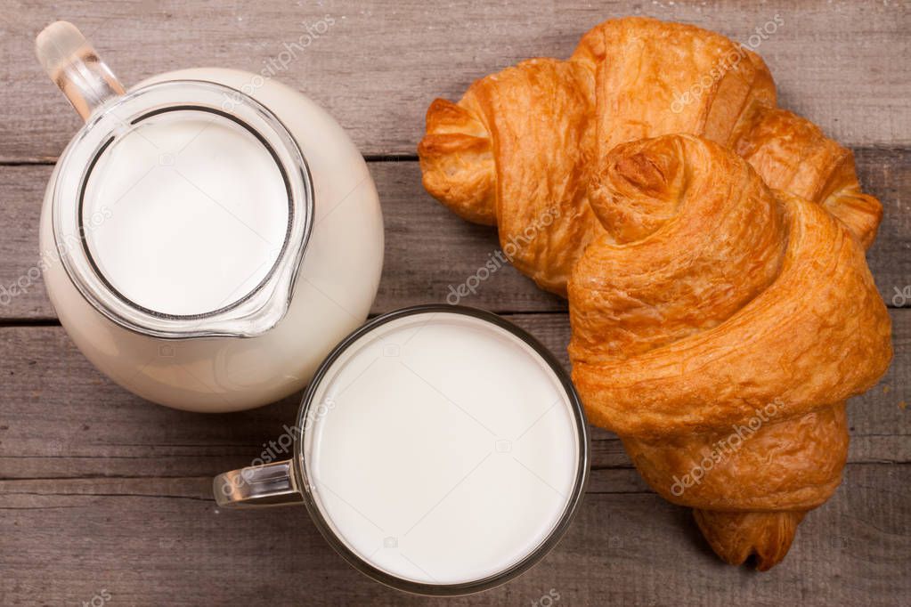 Jug and a glass of milk with a croissant on a wooden background. Top view