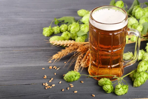 Glass of foamy beer with hop cones and wheat on black wooden background — Stock Photo, Image
