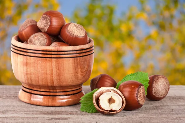 Hazelnuts with leaves in a wooden bowl on a wooden table with blurred garden background — Stock Photo, Image