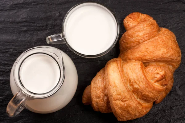 Egy kancsót és egy pohár tej fekete kő alapon két croissant. Szemközti nézet — Stock Fotó