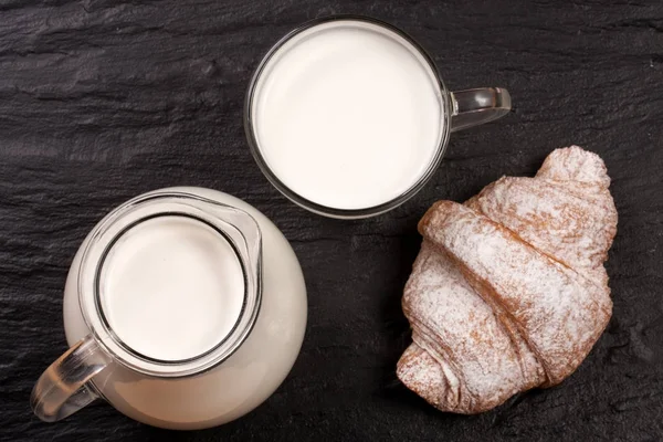 Džbán a sklenice mléka s croissantem na černém pozadí kamene. Pohled shora — Stock fotografie