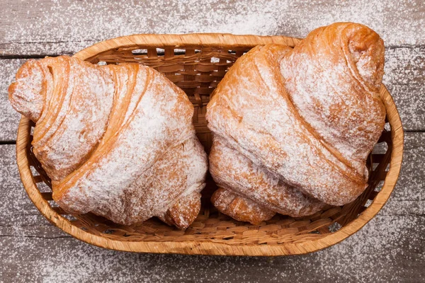 Twee croissants bestrooid met poedersuiker in een rieten mand op oude houten plank bovenaanzicht — Stockfoto