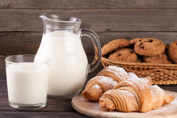 Kancsó és pohár tejet, croissant és zabliszt cookie-kat egy fonott kosárban egy fából készült háttér — Stock Fotó