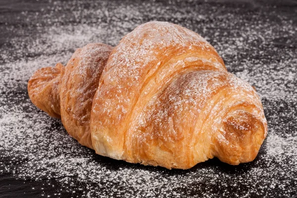 Um croissant polvilhado com açúcar em pó no fundo de pedra preta close-up — Fotografia de Stock