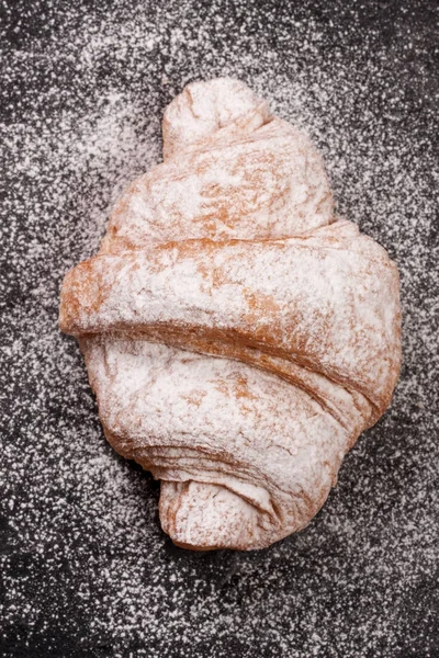 En croissant beströdda med florsocker på svart sten bakgrund närbild. Ovanifrån — Stockfoto