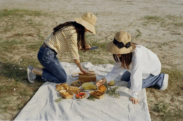 two girls sitting on a picnic table with a basket of wine and a knife
