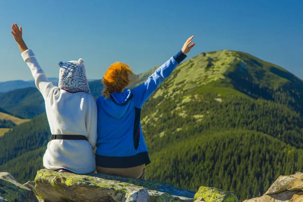 travel photography concept of two girls back to camera sitting on edge of cliff and waving hands on majestic highland green pine forest mountain scenery landscape background in clear bright day time