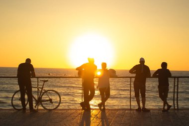 Kameraya geri dönen insanlar, Akdeniz 'in yukarısındaki Tel Aviv rıhtımındaki parlak turuncu gün batımının tadını çıkarıyorlar. Boş fotokopi alanı olan cennet gibi bir arka plan manzarası var. 