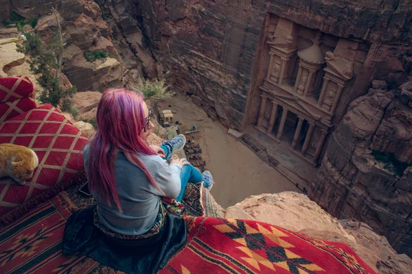 travel photography of lonely girl sitting back to camera on the edge of cliff with top view on Petra ancient treasure exterior building carved in sand stone rock Middle East Jordan tourism destination