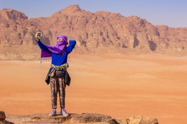 Wadi Rum Ürdün Ortadoğu seyahat fotoğrafçısı tesettürlü Müslüman geleneksel giysiler içinde selfie çeken genç kız muhteşem çöl vadisinde uçurumun kenarında kalıyor ve büyük dağlar var. 