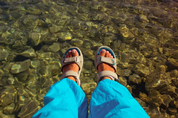 sitting guy male feet in blue sandals and blue pants above lake green shallow water background