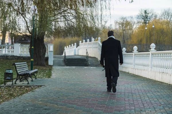 Sonbahar mevsimi sabah vakti siyah ceketli bir adam kameraya geri döner işe gider parka gider banka ve çıplak ağaçlar boyunca asfaltlı yol, kasvetli depresyon atmosferi.