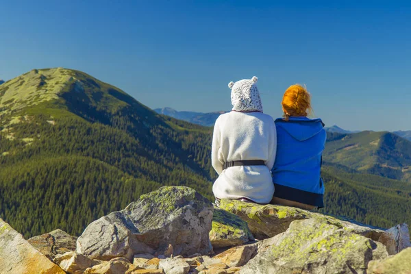 Viajar caminhadas estilo de vida fotografia de duas meninas sentadas de volta à câmera na borda do penhasco de rocha e desfrutar de olhar na paisagem pitoresca vista panorâmica verde montanha espaço de fundo sem foco — Fotografia de Stock