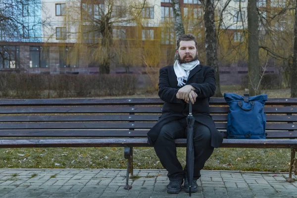 gray depression colors outdoor environment middle adult beard man sitting on a park bench with umbrella in black coat and looking at camera calm without emotions