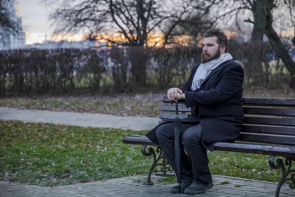 depressive concept photography of single bearded middle adult man portrait sit on a bench in black coat with umbrella and looking side ways in evening park with bare branches