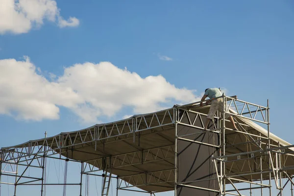 music stage editing by installer worker person back to camera in steel construction shape advertising poster foreshortening from below on blue sky background space for copy or your text here