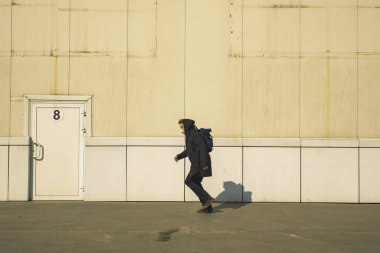 Elinde enfeksiyon kapma riskinden kaçan siyah ceketli bekar bir adam endüstriyel fabrika maskesiyle karşı karşıya. Beyaz dış duvarlı, kapısı kirlenmiş bir çevre alanı.