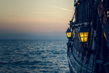 Eski ahşap korsan gemisi günbatımında deniz suyu yüzeyinde romantik zaman diliminde güvertedeki yumuşak odak fenerinin sarı ışığıyla