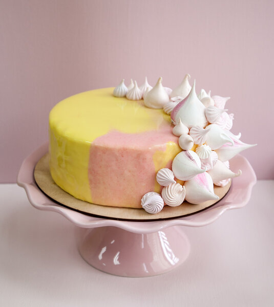 Pink mousse cake covered with yellow and pink chocolate mirror glaze and meringue kisses. On a pink porcelain stand. Pink background. Natural light.