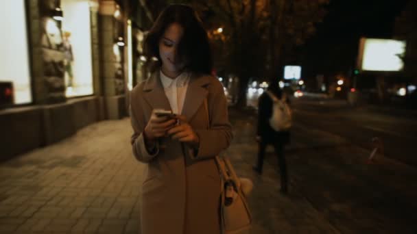 Mujer caminando en la ciudad nocturna mirando hacia abajo a un teléfono inteligente — Vídeos de Stock