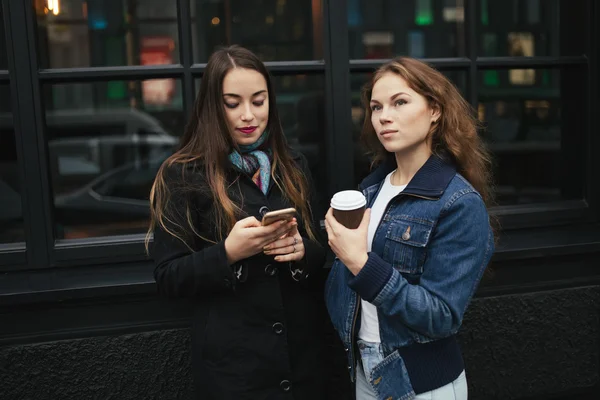 Outdoors portrait of two women drinking coffee and looking at smartphone in city — Stock Photo, Image