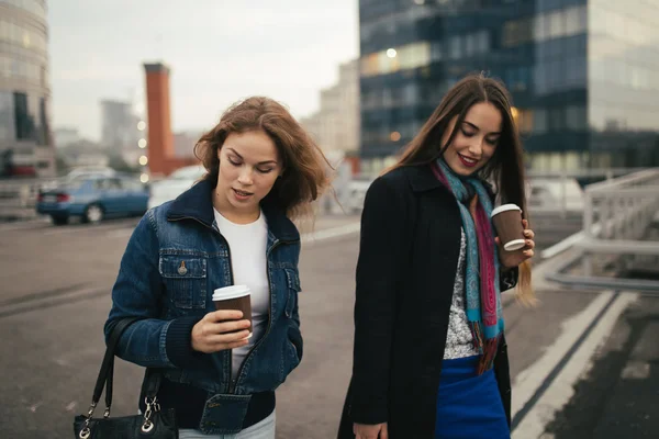 Outdoors fashion portrait of two cheerful girls drinking coffee. Walking in the city. — Stock Photo, Image