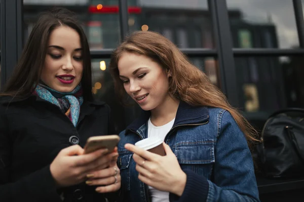 Outdoors portrait of two women drinking coffee and looking at smartphone in city — Stock Photo, Image