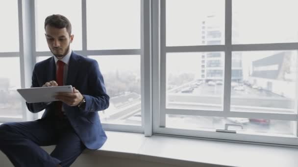 Бизнесмен в белом офисе с помощью цифрового планшета сидит на подоконнике — стоковое видео