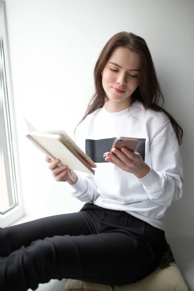 Nuori nainen istuu kannettavan ja älypuhelimen kanssa ikkunalaudalla — kuvapankkivalokuva