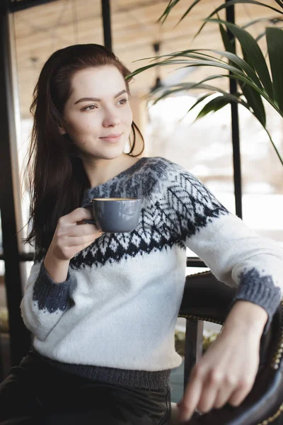 En kvinne i pullover som sitter med en kopp kaffe på en kafe og ser på vinduet – stockfoto