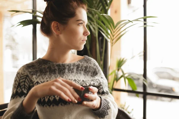 Nainen villapaita pitää kuppi ja katselee ikkunaa kahvilassa — kuvapankkivalokuva