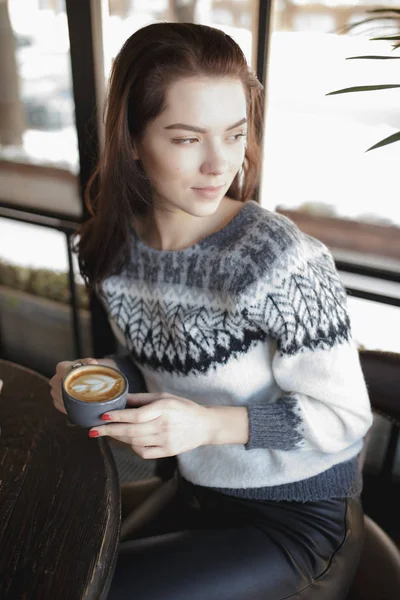 En kvinne i pullover som sitter med en kopp kaffe på en kafe og ser på vinduet – stockfoto
