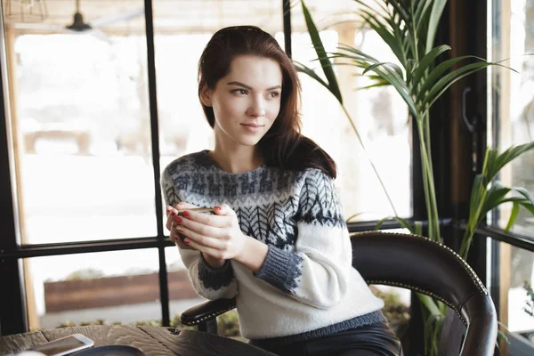ผู้หญิงในชุด pullover นั่งกับถ้วยกาแฟในคาเฟ่และมองไปที่หน้าต่าง — ภาพถ่ายสต็อก
