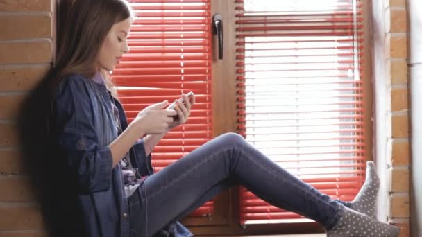 Adolescente chica usando su teléfono inteligente sentado en el alféizar de la ventana — Vídeos de Stock