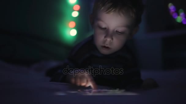 Маленький мальчик играет с планшетным ПК или смартфоном на кровати ночью — стоковое видео